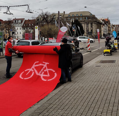 Roter Teppich mit Radwegzeichen wird über ein Auto gerollt, das auf dem Radweg parkt