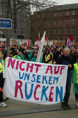 Transparent: Nicht auf unserem Rücken bei Protest gegen geplanten Stellenabbau an Uniklinik Freiburg, am 28.02.2026 am Platz der alten Synagoge; dahinter u.a. eine überdimensionierte Finanzsspritze