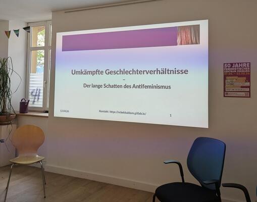 Auf einer weißen Wand mit einem Plakat zum Jubiläum des Feministischen Zentrum Freiburg wird eine Folie projeziert mit dem Titel der Veranstaltung. Vorne sind zwei Stühle zu sehen.