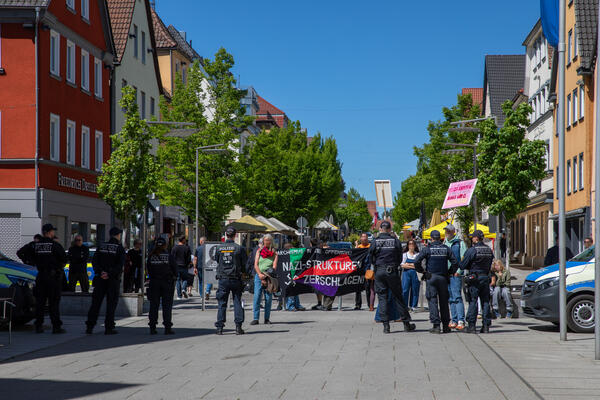 Hinter einer Polizeireihe stehen ein paar antifaschistische Gegendemonstrant*innen mit einem Transparent auf dem "Nazi-Strukturen zerschlagen" steht.