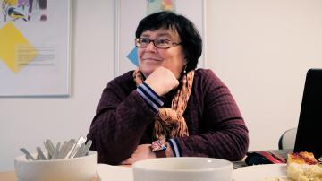 Doritta trägt eine Brille, hat halblanges schwarzes Haar. Sie sitzt am Tisch und lächelt verhalten.