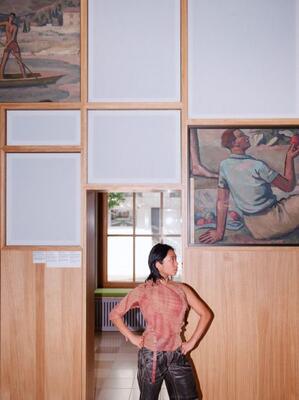 Fassade Jana Baldovino (c) Philip Frowein Schauspielerin Jana Baldovino hat die Hände in die Hüften gestützt + posiert so vor dem faschistischen Wandgemälde von Theodor Kammerer im NS-Dokuzentrum Freiburg + stellt Personen dar,die durch die Wandvertäfelung verdeckt sind.Gemälde ist tlw. sichtbar.