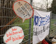 Zwei Schilder mit "Nieder mit der Festung Europa!" und "Wer bleiben will soll bleiben!" sind übereinander in einen Zaun gesteckt. Daneben hängt ein Transparent: "Not safe @ALL - Protection for ALL refugees"