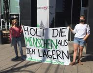 Zwei Frauen halten ein Transparent mit der Aufschrift "Holt die Menschen aus den Lagern" hoch. Sie tragen Mundschutz.