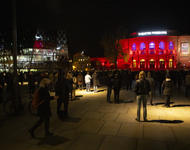 Das Freiburger Stadttheater rot beleuchtet, im Dunkeln, Auf dem Platz davor verteilt stehen Menschen mit Instrumenten.