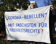 Ein weißes Transparent wird gehalten, auf dem mit blauer Schrift steht: "Corona-Rebellen - Mit Faschisten für Freiheitsrechte?"