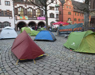 Ein paar Zelte stehen vor einem "Seebrücke jetzt!"-Transparent auf dem Freiburger Rathausplatz. Im Hintergrund sieht man Menschen, einen Pavillion und das Alte Rathausgebäude.