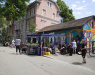 Vor dem kleinen, bunten Haus der Gartenstraße19 steht ein blauer Pavillon und Menschen stehen herum. oder sitzen. Eine Person spricht in ein Mikrofon.