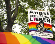 Hinter einem regenbogenfarbenen Pavillondach hängt ein senkrechtes Transparent in den Bäumen auf dem steht: Angst erdrückt das Lieben - united against trans* & homophobia.