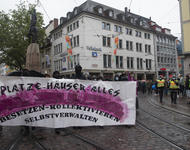 Demonstrationsfront mit einem weißen Transparent auf dem steht: "Plätze, Häuser, alles - Besetzen, kollektivieren, Selbstverwalten." In der Mitte des Transparentes sind pinke Wägen in einem Bogen angeordnet.