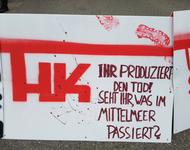Ein weißes Schild mit symbolischen Blutflecken & mit roter Farbe steht geschrieben: "HK - Ihr produziert den Tod! Seht ihr, was im Mittelmeer passiert?"