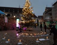 Viele brennende Kerzen stehen auf dem Ni-Una-Menos-Platz vor der bunt leuchtenden Säule der Toleranz und einem beleuchteten Tannenbaum. An der Wand hängt eine Trans-Flagge.