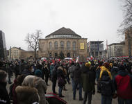 Platz der Alten Synagoge mit Blick auf das Stadttheater. Menschenmenge mit Fahnen und Schildern. Auf der rechten Seite des Theaters ist ein gelbes Plakat angebracht: "Impfen schützt Leben und die Kultur"