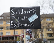 Ein Kind, das dick angezogen ist und eine medizinische Maske trägt hält ein schwarzes Schild in die Höhe auf dem in weiß "Solidarität statt Egoismus" steht. Eine medizinische Maske und eine Spritze sind aufgeklebt.