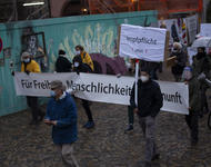Eine Person in dunkler, dicken Jacke und in grauer Hose, trägt vor einer Querdenken-Demonstration ein Schild, auf dem "Impfpflicht jetzt!" steht.