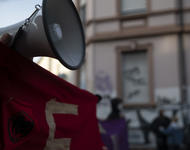 Ein Megafon ragt über ein rotes Transparent in der Nahaufnahme. An dem Transparent ist ein roter Aufnäher mit schwarzem Antifa-Logo befestigt. Mit Sicherheitsnadeln.