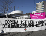 Transparent auf dem Europaplatz. Auf weißem Stoff steht "Corona ist der Virus - Kapitalismus ist die Krise". Auf der Unterseite ist mittig ein Antifa-Logo angeordnet. Hinder dem Transparent sieht man ein pinkes Hochtransparent, mit dem Demomotto.