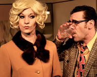 Die Schauspielerin Chris Meiser (Rosemarie) steht mit blonder Perücke und beigem Kostüm neben ihrem Spielpartner Jochen Kruß, der im Karierten Jackett und bunter Krawatte ein Glas auf Ex trinkt G