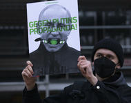 Ein männlich gelesener Demonstrant mit schwarzer Mütze wie Maske hält ein Schild in die Höhe, auf dem Putin mit einem Knebel abgebildet ist. Es steht "Gegen Putins Propaganda!" darauf.