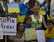 Ausschnitt aus einer Formation Ukrainischer Demonstrierender. Auf der rechten Bildhälfte eine Frau mit zwei Kindern an ihren beiden Händen, alle in der Hocke. Linkes Kind mit Schild mit gelb-blauem Davidstern, rechtes 1 auf dem steht: We want to go home.