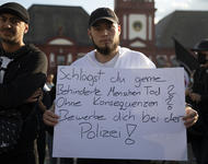 Portraitaufnahme: Zwei männliche Personen stehen auf dem Marktplatz in Mannheim nebeneinander. Die Person im Zentrum trägt ein weißes Schild auf Brusthöhe: "Schlägst du gerne Behinderte Menschen Tod? Ohne Konsequenzen? Bewerbe dich bei der Polizei!"