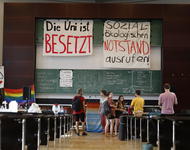 Zentrierter Blick in einen Hörsaal. Hinten ist das Redner*innen-Pult zu sehen, ein paar Menschen und zwei Banner die hoch oben an einer Tafel hängen. "Die Uni ist besetzt" und "Sozial-ökologischen Notstand ausrufen" steht darauf.