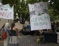 Es sind zwei Menschen vor an zwei auf einer Schnur befestigten Transparenten zu sehen. Auf dem linken Transpi steht "Wir campen bis ihr handelt!", auf dem Anderen: "Klimacamp Freiburg" mit einer Sillhouette der Blauen Brücke und drei Zelten darauf.