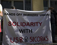 Zwei Personen, halten und befinden sich hinter einem weißen Transparent auf dem steht in schwarz und rot: Hands off workerss unions - Solidarity with USB & SICOBAS. 