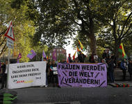 Demonstrant*innen am Platz der Alten Synagoge in Freiburg auf der Seite zur Bertoldstraße. Es sind kurdische Fahnen zu sehen und zwei Transparente. Auf dem rechten steht: "Frauen werden die Welt verändern!", in Großbuchstaben, auf violettem Stoff.
