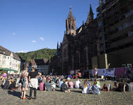 Blick auf dem Münsterplatz mit dem hinteren Teil des Münsters im Hintergrund. Rechts sind Transparente und eine kleine Bühne zu erkennen. Vorne sitzende und paar stehende Menschen, die einer redenden Person zuhören.