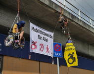 3 Menschen, zwei davon im Rollstuhl hängen kletternd vor dem Bahnhof Frankfurt West mit großen Bannern, die für eine Mobilitätswende für Alle werben.