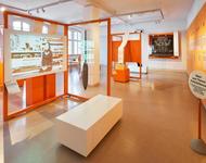  Blick in einen in Orange getauchten Ausstellungsraum gefüllt mit Vitrinen und Projektionsflächen, auf denen Filme laufen 