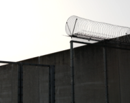 Hohes Gittertor, rechts etwas höher mit spiralförmigem Stacheldraht. Dahiner Gefängnismauer.