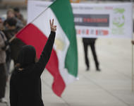 Eine Person mit langen braunen haaren, schwarzer Filzmütze und schwarzer Kleidung ist von hinten zu sehen, wie sie ihre rechte Hand zum Victory & Peace - Zeichen in die Höhe streckt. Im Hintergrund unscharf eine Iran-Fahne zu sehen und weitere Menschen.