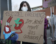 Eine Person mit weißer FFP2-Maske und langen braunen Haaren hält das Schild mit Mr. Crabs von Spongebob Schwammkopf, der ein Geldbündel in der Hand hält: "1,5°C-Ziel erreichen? 'Der Markt regelt das schon!'"