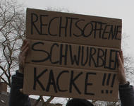 Ein Pappschild auf dem "Rechtsoffene Schwurblerkacke!!!" steht wird mit zwei Händen in die Höhe gehalten. Im Hintergrund grauer Himmel und blattlose Baumkronen zu sehen.