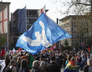 Im Fokus des Bildes befindet sich eine blau-weiße Friedenstauben-Fahne. Es sind Demonstrant*innen auf dem Platz der Alten Synagoge zu sehen.