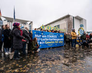 Eine Gruppe Menschen steht mit Banner vor dem Bundeskanzleramt. SChriftzug: Nur ein starkes Lieferkettengesetz ist ein Gewinn für alle. Darüber die Zahl 90.248 der geleisteten Unterschrifen der Petition.