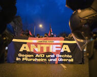 Eingerahmt von Helm und Schusswaffe von Polizei ist das Fronttransparent einer Demonstration zu erkennen, und ein paar rote Fahnen. Es steht darauf "ANTIFA - Gegen AfD und Rechte in Pforzheim und überall".