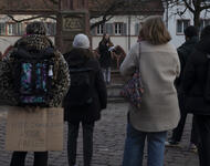 Vier Menschen stehen mit dem Rücken zur Kamera im Bild. Person links trägt Leojacke und ein Pappschild vor dem Gesäß: 10 Jahre ich hier immer noch keine Papiere. Im Hintergrund Rednerin vor dem Brunnen auf dem Rathausplatz.