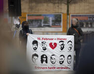 Zwei Personen halten ein Transparent mit neun aufgemalten Portraits der Getöteten. Oben in rot mit einem zerbrochenem Herzen "19. Februar - Hanau", unten: "Erinnern heißt verändern."