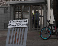Das Eingangstor der LEA Freiburg im Hochformat. Vorne ein Schild mit Bleiberecht statt Abschiebeknast. Hinten das offizielle Schild und ein*e Bewohner*in, die gerade von Security durchsucht wird.