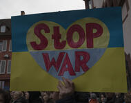 Ein Schild mit den Farben der Ukraine wird ins Bild gehalten. Ein Herz ist abgebildet, mit invertierten Faben (gelb-blau) und darauf die Worte: Stop War