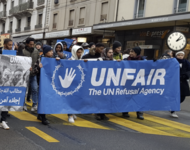 Demonstrierende auf der Straße mit blauem Banner: UNFAIR - The UN Refusal Agency