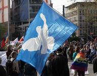 Eine blaue Fahne mit weißer Friedenstaube ist in Bildmitte zu sehen. Dahinter bzw. daneben ein Demonstrationspublikum.