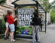 Eine Gruppe Menschen hängt ein Plakat in ein Bushäuschen im Camouflage-Stil der Bundeswehr: "KEIN Diensta an der Waffe geht OHNE Waffe"