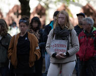 Mehrere Menschen stehen in Jacken gekleidet nebeneinander. Eine Person mit langen, blonden Haaren hält ein Schild vor ihren Bauch, auf dem steht: "Stopt das Sterben an unseren Grenzen"