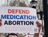 Ein Plakat wird von einer Person hochgehalten, die nicht im Bild ist: Defend Medication Abortion. Bigger than Roe
