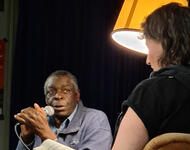 David Macou im Gespräch mit Kathi King in der Mensabar. Sie sitzen in Sesseln, sprechen ins Mikro. Zwischen ihnen eine Wohnzimmerlampe