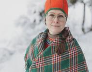 Åsa Larsson Blind in traditioneller Sámikleidung mit grün-rot karierter Decke umgeschlungen und orangener Mütze vor einer schneebedeckten Landschaft